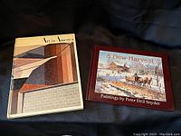 Two hardcover art books side by side on a black fabric background. Left: "Art in America" with abstract architectural cover art. Right: "A New Harvest" with a winter rural painting cover, burgundy background.