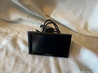 Front view of Garmin GPS device with screen visible and Garmin logo on the bottom bezel. Car charger cable visible behind device.
