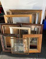 Front view of 10 assorted picture frames stacked leaning against a white wall showing variety in size, wood tones, and styles.