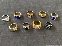 All 9 vintage costume rings displayed together on dark fabric background showing various sizes, stone colors and metals.