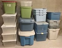 Array of twenty large plastic storage tubs in different colors and sizes, stacked and arranged on a cement floor, some with lids and some without.