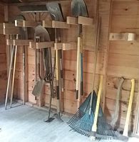 Garden tools mounted on wooden wall including shovels, rakes, axes and hoes in a shed.