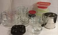 Photo showing a variety of clear glass Pyrex bowls and pie plates, several plastic bowl covers in red and green, a metal large flour sifter, clear glass measuring cups with red measurements, and a black silicone 6-cup egg cooker.