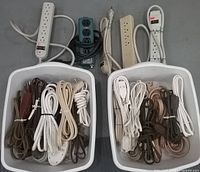 Two white bins filled with assorted indoor electrical cords in white, brown, beige, and black, along with six different power strips of various lengths and outlet counts.