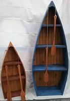 Photo showing two wooden canoe-shaped shelves side by side, one large blue painted and one smaller pink stained. Both have multiple horizontal shelves inside and two wooden paddles hanging by strings.