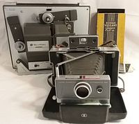 Photo showing three vintage photography items: Polaroid camera in front, Bell & Howard super 8 projector behind it, and Kodak Vigilant box to side with paperwork.