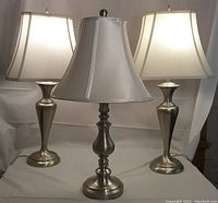 Photo showing the set of three table lamps with silver metal bases and white fabric shades. Two lamps are 27 inches tall, the third lamp is slightly shorter at 25 inches.