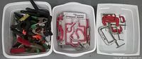 Three white bins containing assorted tools: first bin has various plastic spring clamps in multiple colors; second bin has packaged assorted storage hooks plus loose hooks; third bin has red wooden C-clamps and metal clamps with adjustable screws.