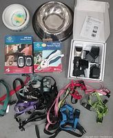 Full overview of the lot showing the electronic dog trainers in their packages, dog clipper with accessories in box, assorted colorful small dog leashes and harnesses, one ceramic dog bowl and one stainless steel dog bowl.
