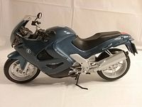 Side view of large 1:6 scale BMW motorcycle model with metallic blue fairing, black seat, silver wheels and exhaust. Model rests on kickstand showing details of tires, foot pegs, and handlebar controls.