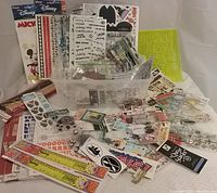 Overall view of assortment of over 70 packages of scrapbooking stickers, stencils, rulers, and accents arranged with a plastic container holding some items.