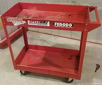 Red metal shop cart with two shelves, red paint showing signs of use and dirt, four caster wheels, and three racing brand stickers on one side.