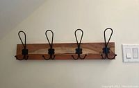 Wall-mounted wood backed coat rack with four black metal hooks against a light wall background.