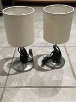 Photo showing two identical desk lamps with white cylindrical shades and metal bases placed on tiled floor with cords coiled around bases.