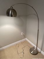 Full view of the adjustable silver floor lamp standing on carpet against a wall, plugged in with visible cord and foot switch.