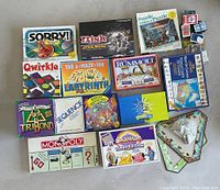 Photo showing assorted classic board games and puzzle boxes arranged on a floor. Visible game titles include Sorry!, Risk Star Wars, Qwirkle, Labyrinth, Rummoli, Settlers of Catan, TriBond, Sequence, Pictionary, Scrabble, Monopoly, Cranium Turbo Edition. Also includes jigsaw puzzles and a deck of cards.