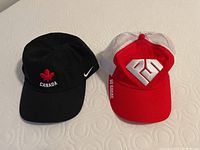 Two baseball style hats: black Nike cap with Canada maple leaf, red and white trucker hat with 'BE GREAT' branding.