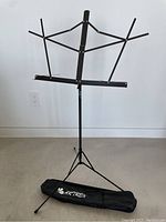 Black metal height adjustable music stand set up with tripod base next to its black carry case lying flat on the floor