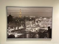 Large black and white framed photograph showing panoramic view of Paris cityscape focused on Eiffel Tower and river bridges at night with city lights.