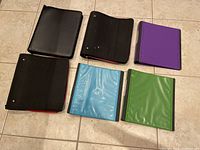 Six hard cover school binders in black, purple, blue, green laid out on floor, showing three with zipper closures and three without.