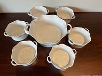 All seven white ceramic bowls including one large casserole bowl and six smaller matching bowls, displayed on a wooden surface