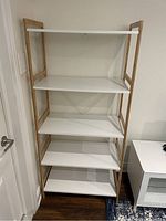 Front view of the white shelving unit with light brown frame against a wall showing the four shelves and open side design.