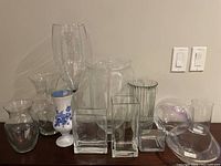 Wide photo showing all 11 glass vases arranged on a wooden surface against a beige wall.