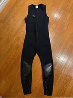 Full-length front view of black sleeveless wetsuit laid flat on wooden floor showing front zipper and knee pad reinforcement.
