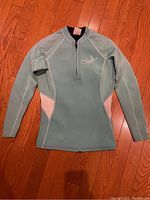 Front view of a light bluish-green O'Neill wet suit top with long sleeves on wooden floor