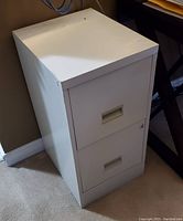 Front and side view of white 2 drawer steel file cabinet with recessed handles, placed in corner of room on carpeted floor.