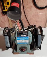 Front view of Delta 6-inch bench grinder showing coarse and fine grinding wheels with spark shields, central power switch, and attached goose neck light above the grinder.