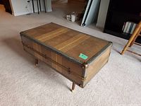 View of the rectangular antique suitcase coffee table on four legs from an angle, showing its brown leather exterior with lighter horizontal stripes and metal studs.