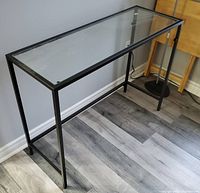 Side angle view of black metal frame side table with clear rectangular glass top on grey wood effect floor.