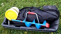 Photo of manual ice auger, yellow lid minnow bucket, black carrying bag likely containing ice fishing tent, all placed on a black plastic toboggan sled sitting on grass.