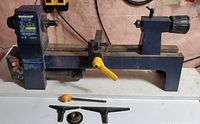 Full view of Mastercraft Mini Wood Lathe mounted on wooden base with yellow adjustment lever and accessories in front.
