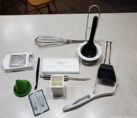 Photo shows 13 various kitchen utensils arranged on a countertop, mostly white plastic items with some metal and black accents, including a whisk, peelers, tongs, and specialty tools.