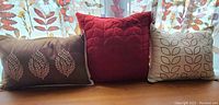 Photo shows three decorative pillows arranged on a wooden surface by a window with sheer floral curtains. Pillows have fall leaf patterns in various colors and sizes.