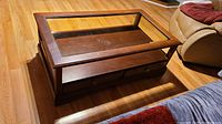 Wooden coffee table showing the inset glass top, wooden frame, and drawer front with metal knob. The table is on a wooden floor next to a sofa.