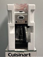 Front view of the coffee maker partially encased in white protective Styrofoam packaging with glass carafe visible.