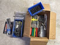 Overall view of a large cardboard box and blue plastic bins containing numerous loose metal drill bits and some packaged drill accessories.