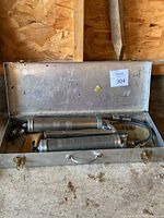Open metal carrying case containing two metal grease dispensers with hoses and lever handles. Case shows dirt and wear.