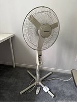 Full view of white Honeywell pedestal fan with protective mesh grill and three blades. Fan is standing on carpet with four legs for stability. Power cord visible and remote control laying on floor near stand.