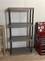 Full view of gray plastic utility shelf with four shelves standing against wall next to a red toolbox.