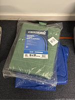 Photo shows two tarps stacked: a green Certified brand tarp 15x20 feet in factory packaging on top of a blue used tarp, both made of polyethylene.