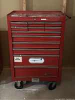 Front view of red Mastercraft rolling toolbox showing all 12 closed drawers, caster wheels, scratches and wear on top surface, and various stickers on front.