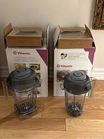 Two Vitamix blender containers with handles and lids placed in front of corresponding Vitamix product boxes.