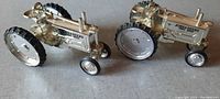 Two chrome-colored John Deere toy tractors with narrow wheels placed on a gray surface, measurements approximately 3 x 3 inches, showing left and right side views.