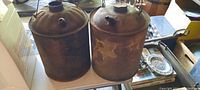 Two old, rusty gallon-sized fuel cans with round screw top caps and small vent spouts, displayed side by side on a table with visible barn dust and wear.