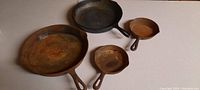 Four cast iron frying pans displayed on a light surface. Large rusty 12 inch pan, slightly less rusty 10 inch pan, and two smaller 6 inch pans, one marked Griswold.