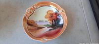 Top-down view of the serving dish showing the detailed handpainted landscape and the three small handles spaced around the rim.